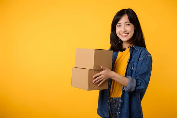 stock image Portrait young asian woman wearing yellow t-shirt and denim shirt holding parcel box isolated on yellow studio background, Delivery courier and shipping service concept.