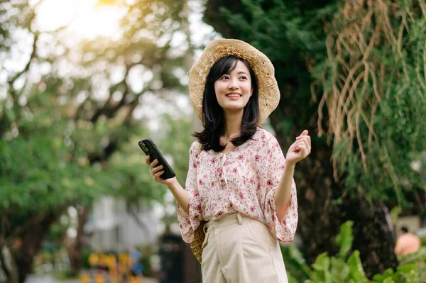 stock image Portrait of young asian woman traveler with weaving hat and basket using mobile phone on green park nature background. Journey trip lifestyle, world travel explorer or Asia summer tourism concept.