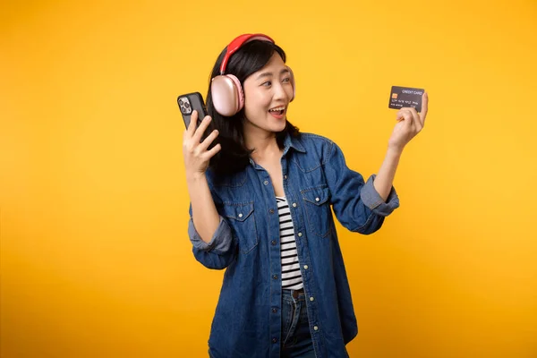 デニムの服を着た笑顔とスマートフォンやクレジットカードを持つヘッドフォンを身に着けて幸せな成功を収めた肖像アジアの若い女性 ショッピングオンライン携帯電話エンターテイメントライフスタイルコンセプト — ストック写真