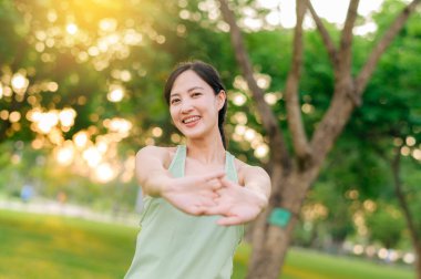 Kadın koşucu. Açık havada koşup eğlenmeden önce yeşil spor kıyafetli sağlıklı bir Asyalı genç kadın parkta kaslarını esnetiyor. Kamu parkındaki spor koşucusu kız. Refah konsept demek.