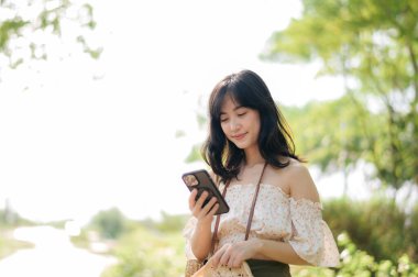 Yeşil park arka planında şapkası, sepeti, cep telefonu ve kamerası olan Asyalı genç bir kadın gezginin portresi. Seyahat yaşam tarzı, dünya turisti ya da Asya yaz turizmi konsepti.