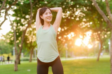 Kadın koşucu. Açık havada koşup eğlenmeden önce yeşil spor kıyafetli sağlıklı bir Asyalı genç kadın parkta kaslarını esnetiyor. Kamu parkındaki spor koşucusu kız. Refah konsept demek.