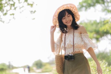 Dokuma şapkası ve sepeti ve yeşil halka açık park arka planında kamerası olan Asyalı genç bir kadın gezginin portresi. Seyahat yaşam tarzı, dünya turisti ya da Asya yaz turizmi konsepti.