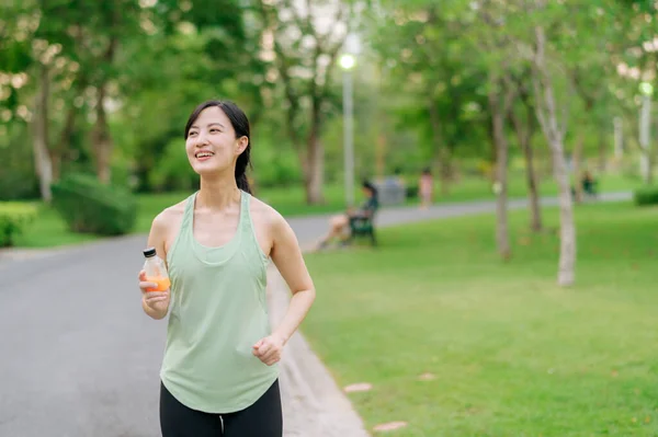 Female jogger. Fit Asian young woman with green sportswear drinking organic orange juice after running and enjoying a healthy outdoor. Fitness runner girl in public park. Wellness being concept