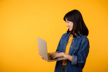 Sarı tişört ve kot gömlek giyen, dizüstü bilgisayar tutan mutlu bir genç kadın portresi ve sarı stüdyo arka planında izole edilmiş not defteri. iş teknolojisi uygulama iletişim kavramı.