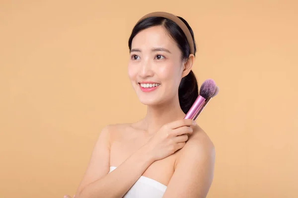 Portrait young happy asian woman with natural make up face holding cosmetic skin powder blusher isolated on beige background. Female apply skincare brush treatment. beauty product, cosmetology concept.