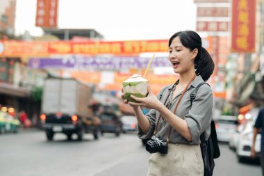 Happy young Asian woman backpack traveler drinking a coconut juice at China town street food market in Bangkok, Thailand.