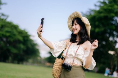 Yeşil park arka planında şapkası, sepeti, cep telefonu ve kamerası olan Asyalı genç bir kadın gezginin portresi. Seyahat yaşam tarzı, dünya turisti ya da Asya yaz turizmi konsepti.