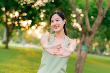 Kadın koşucu. Açık havada koşup eğlenmeden önce yeşil spor kıyafetli sağlıklı bir Asyalı genç kadın parkta kaslarını esnetiyor. Kamu parkındaki spor koşucusu kız. Refah konsept demek.