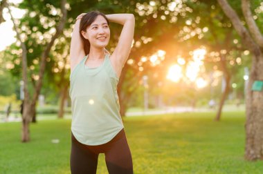 Kadın koşucu. Açık havada koşup eğlenmeden önce yeşil spor kıyafetli sağlıklı bir Asyalı genç kadın parkta kaslarını esnetiyor. Kamu parkındaki spor koşucusu kız. Refah konsept demek.