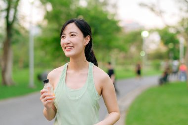 Kadın koşucu. Yeşil spor kıyafetli, sağlıklı bir açık havada koşup eğlendikten sonra organik portakal suyu içen formda bir Asyalı genç kadın. Kamu parkındaki spor koşucusu kız. Refah konsept demek.