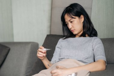 Kanepede öksürürken klinik termometre okuyan hasta Asyalı kadın..