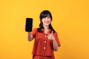 Portre güzel Asyalı kadın turuncu elbiseli mutlu bir gülümseme sarı stüdyo arka planında izole edilmiş akıllı telefona el hareketini işaret ediyor. uygulama akıllı telefon kavramı