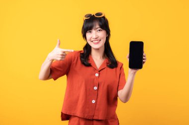 Portre güzel Asyalı kadın turuncu elbiseli mutlu bir gülümseme sarı stüdyo arka planında izole edilmiş akıllı telefona el hareketini işaret ediyor. uygulama akıllı telefon kavramı