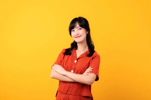 穿着橙色衣服的年轻而体贴的美丽的亚洲女人快乐地微笑着 在黄色的摄影棚背景上表现出交叉手臂的动作 — 图库照片