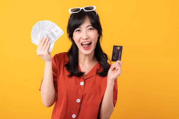 年轻美丽的亚洲女人 穿着橙色衣服 露出现金和信用卡 背景黄色 面带微笑 容光焕发 付款及购物付款概念 — 图库照片