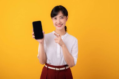 Genç, güzel Asyalı girişimci beyaz gömlek ve kırmızı bitkiler giymiş mutlu bir gülümseme takınarak sarı arka plandaki akıllı telefon izolasyonuna parmak işareti yapan portre. Yeni uygulama kavramı sunuluyor