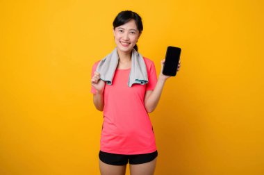 Asyalı genç sporcu kadın mutlu bir gülümseme, pembe spor giyim ve akıllı telefon, sarı stüdyo arka planında egzersiz yapıyor. Teknoloji sağlık yaşam tarzı kavramı.