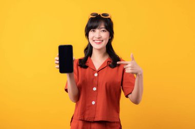 Portre güzel Asyalı kadın turuncu elbiseli mutlu bir gülümseme sarı stüdyo arka planında izole edilmiş akıllı telefona el hareketini işaret ediyor. uygulama akıllı telefon kavramı