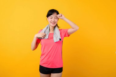 Asyalı genç spor fitness kadını portresi. Pembe spor kıyafetleri ve yüz havlusu giymiş mutlu bir gülümseme. Sarı stüdyo arka planında egzersiz yapıyor. Refah ve sağlıklı yaşam tarzı kavramı.