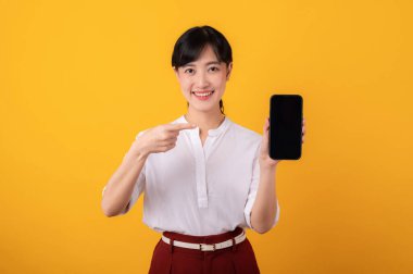 Genç, güzel Asyalı girişimci beyaz gömlek ve kırmızı bitkiler giymiş mutlu bir gülümseme takınarak sarı arka plandaki akıllı telefon izolasyonuna parmak işareti yapan portre. Yeni uygulama kavramı sunuluyor