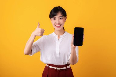 Genç, güzel Asyalı girişimci kadın mutlu bir gülümseme beyaz gömlek ve kırmızı bitkiler giyerek sarı arka plandaki akıllı telefon izolasyonuna işaret ediyor. Yeni uygulama kavramı sunuluyor