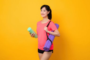 Asyalı genç spor fitness kadını portresi. Pembe spor kıyafetleri ve yoga minderi giymiş mutlu bir gülümseme. Sarı stüdyo arka planında egzersiz yapıyor. Sağlıklı sağlık yaşam tarzı kavramı.