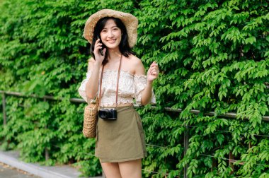 Yeşil park arka planında şapkası, sepeti, cep telefonu ve kamerası olan Asyalı genç bir kadın gezginin portresi. Seyahat yaşam tarzı, dünya turisti ya da Asya yaz turizmi konsepti.