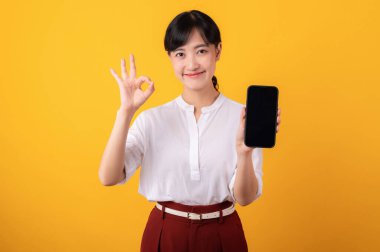 Genç, güzel Asyalı girişimci kadının portresi beyaz gömlek ve kırmızı bitkiler giyerek mutlu bir gülümseme takınıyor ve sarı arka plandaki akıllı telefon izolasyonuna el hareketi yapıyor. Yeni uygulama kavramı sunuluyor