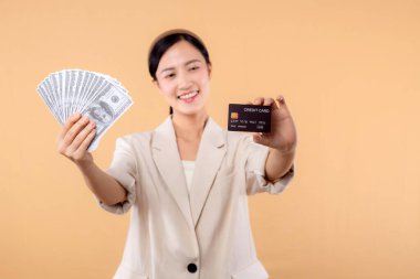 Beyaz ceketli mutlu Asyalı iş kadınının portresi. Elinde nakit para ve kredi kartı var. Bej arka planda duruyor. Milyoner işi, alışveriş konsepti