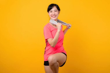 Asyalı genç spor fitness kadını portresi. Pembe spor kıyafetleri ve yüz havlusu giymiş mutlu bir gülümseme. Sarı stüdyo arka planında egzersiz yapıyor. Refah ve sağlıklı yaşam tarzı kavramı.