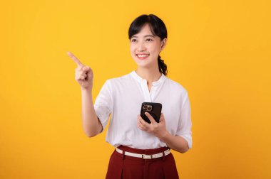 Genç, güzel Asyalı girişimci mutlu bir gülümseme beyaz gömlek ve kırmızı bitkiler giyerek bedava fotokopi alanına parmak işareti yaparak sarı arka planda akıllı telefon izolasyonu kullanarak..