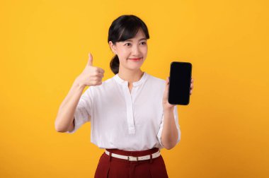 Genç, güzel Asyalı girişimci kadın mutlu bir gülümseme beyaz gömlek ve kırmızı bitkiler giyerek sarı arka plandaki akıllı telefon izolasyonuna işaret ediyor. Yeni uygulama kavramı sunuluyor