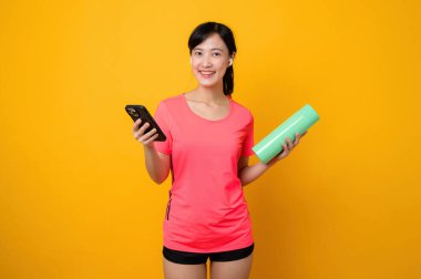 Asyalı genç sporcu kadın mutlu bir gülümseme, pembe spor giyim ve akıllı telefon, sarı stüdyo arka planında egzersiz yapıyor. Teknoloji sağlık yaşam tarzı kavramı.
