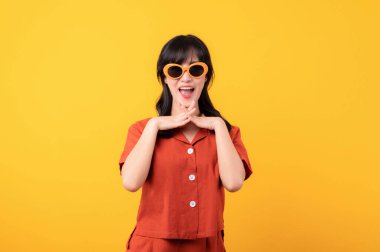 Güzel Asyalı kadın portresi turuncu kıyafetler ve güneş gözlüğü takmış mutlu bir gülümseme. Sarı stüdyo arka planında, yüzünde izole edilmiş bir el ile kutlama yapıyor..