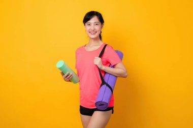 Asyalı genç spor fitness kadını portresi. Pembe spor kıyafetleri ve yoga minderi giymiş mutlu bir gülümseme. Sarı stüdyo arka planında egzersiz yapıyor. Sağlıklı sağlık yaşam tarzı kavramı.
