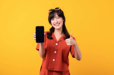 Portre genç, güzel Asyalı kadın mutlu bir gülümseme turuncu kıyafetler giymiş, el hareketi yapıyor ve sarı arka planda izole edilmiş akıllı telefonu tutuyor. uygulama akıllı telefon kavramı