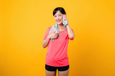 Asyalı genç spor fitness kadını portresi. Pembe spor kıyafetleri ve yüz havlusu giymiş mutlu bir gülümseme. Sarı stüdyo arka planında egzersiz yapıyor. Refah ve sağlıklı yaşam tarzı kavramı.