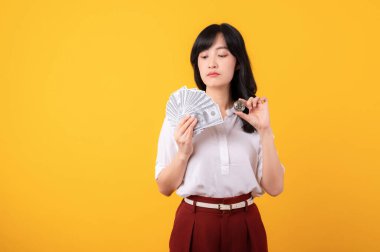 Beyaz gömlek ve kırmızı bitkiler içinde nakit para ve sarı arka planda kripto dijital para birimi izolasyonu tutan şüpheli yüzlü genç Asyalı kadın girişimci portresi. Zenginlik kavramı