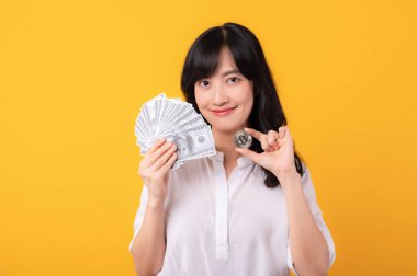 Asyalı genç ve güzel girişimci kadının portresi beyaz gömlek ve kırmızı bitkiler içinde nakit para ve sarı arka planda kripto dijital para birimi izolasyonu tutan mutlu bir gülümseme. Zenginlik kavramı.