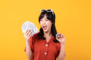 Portre güzel, genç Asyalı mutlu bir gülümseme. Elinde nakit para ve kripto bitcoin dijital para var. Sarı arka planda izole edilmiş başarısını kutluyor. Zengin ol..