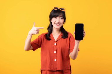 Portre güzel Asyalı kadın turuncu elbiseli mutlu bir gülümseme sarı stüdyo arka planında izole edilmiş akıllı telefona el hareketini işaret ediyor. uygulama akıllı telefon kavramı