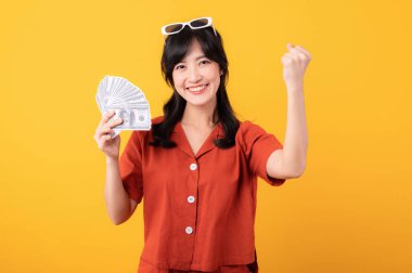 Portre güzel genç Asyalı kadın turuncu giysiler içinde mutlu bir gülümseme nakit para taşıyor ve başarısını sarı arka planda yalıtılmış bir şekilde kutluyor. Zengin ol..