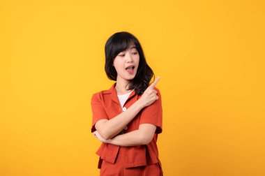 Portre güzel, genç Asyalı mutlu gülümseyen turuncu elbiseli, sarı stüdyo arka planında izole edilmiş boş alana işaret eden el hareketini gösteren bir resim. Güzel ürünlere dikkat.