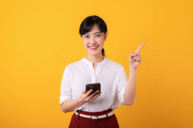 Genç, güzel Asyalı girişimci mutlu bir gülümseme beyaz gömlek ve kırmızı bitkiler giyerek bedava fotokopi alanına parmak işareti yaparak sarı arka planda akıllı telefon izolasyonu kullanarak..