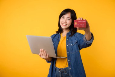 Asya kökenli mutlu genç bir kadın dizüstü bilgisayar kullanarak sarı tişört giyer kredi kartı alışverişi yapar sarı arka planda izole edilmiş banka kartı siparişi verir..