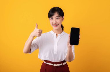 Genç, güzel Asyalı girişimci kadın mutlu bir gülümseme beyaz gömlek ve kırmızı bitkiler giyerek sarı arka plandaki akıllı telefon izolasyonuna işaret ediyor. Yeni uygulama kavramı sunuluyor