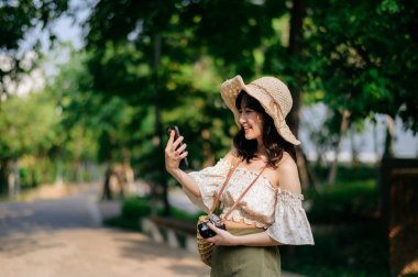 Yeşil park arka planında şapkası, sepeti, cep telefonu ve kamerası olan Asyalı genç bir kadın gezginin portresi. Seyahat yaşam tarzı, dünya turisti ya da Asya yaz turizmi konsepti.