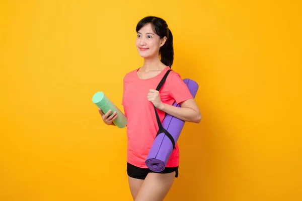 Portrait asian young sports fitness woman happy smile wearing pink sportswear and yoga mat doing exercise training workout against yellow studio background. Healthy wellness lifestyle concept.