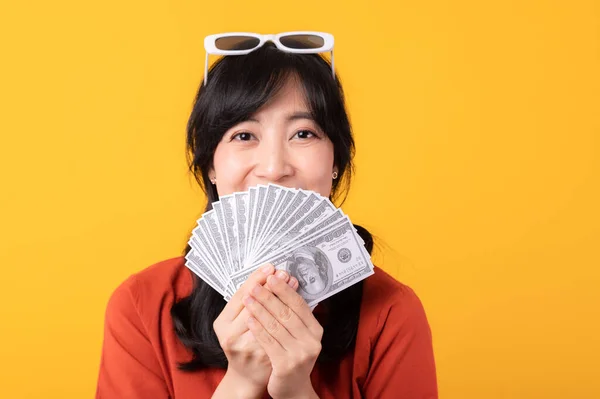 穿着橙色衣服的美丽的年轻亚洲女人 笑容满面 带着钱 背景是黄色的 付款及购物付款概念 — 图库照片